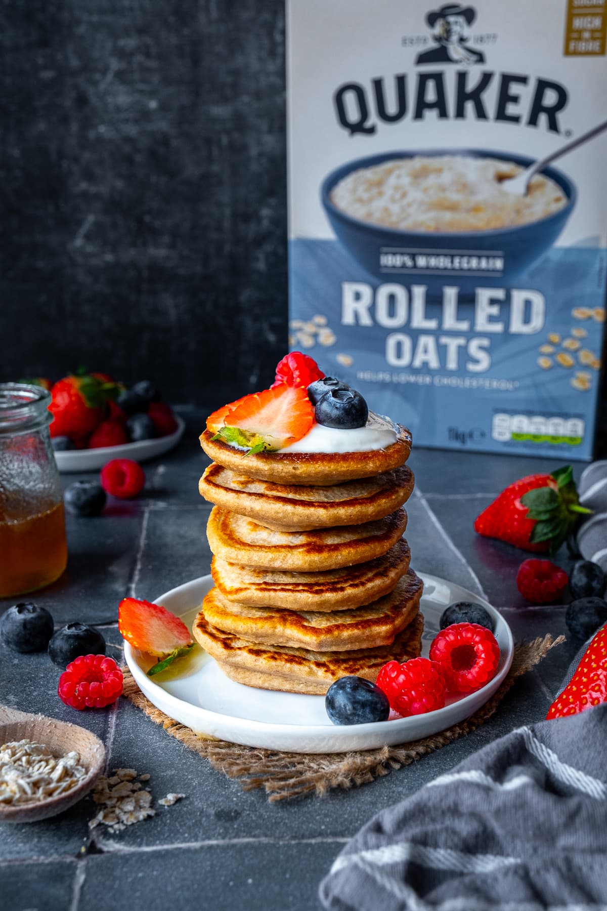 Stack of 6 Banana Oat Egg Pancakes with berries and yogurt, in front of a Quaker Rolled Oats box