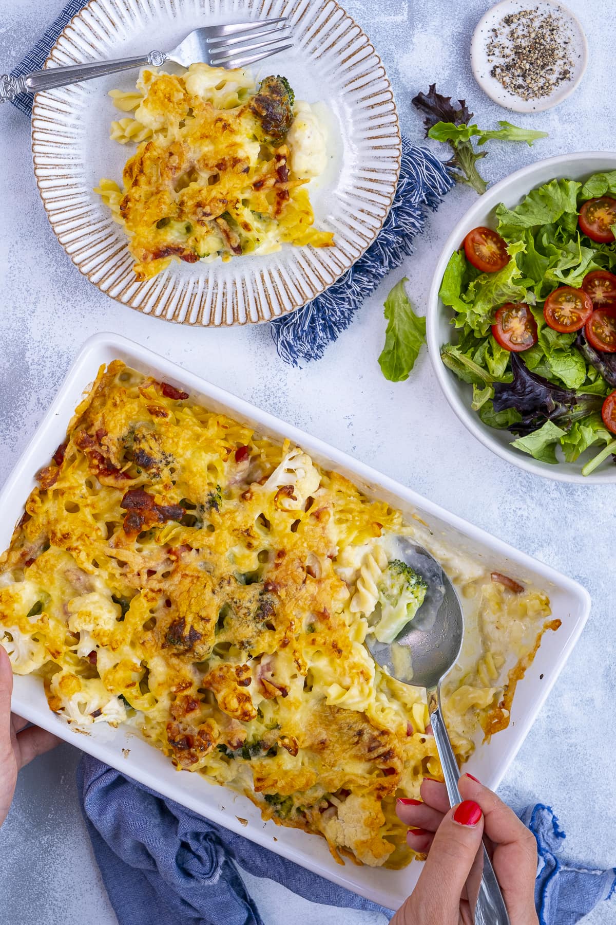 Overhead shot of Cauliflower, Broccoli and Bacon Pasta Bake in an oven dish. One portion is on a plate.