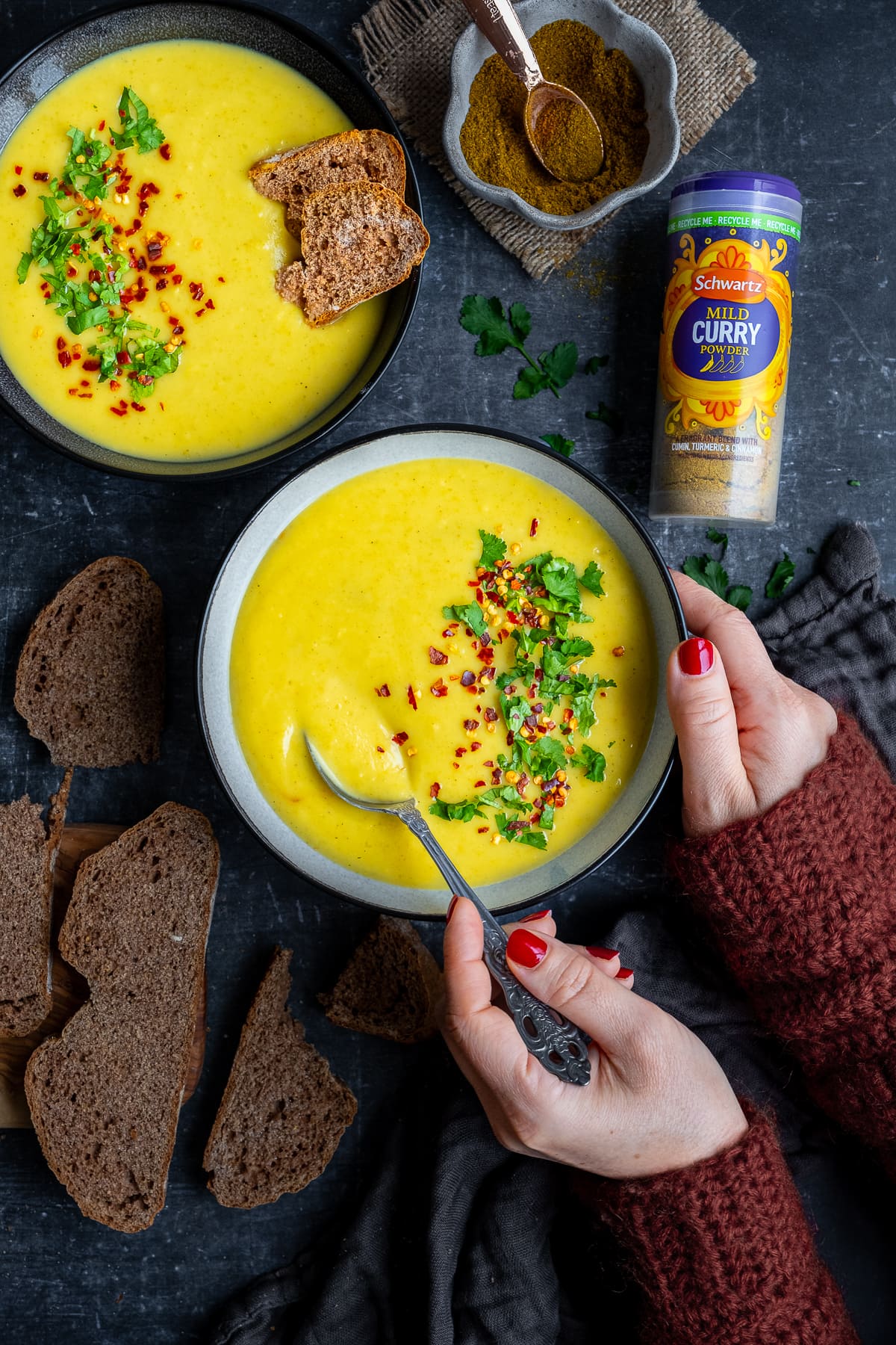 Overhead shot of 2 bowls of Curried Parsnip Soup. A woman's hands holding a spoon, taking a spoonful of soup.