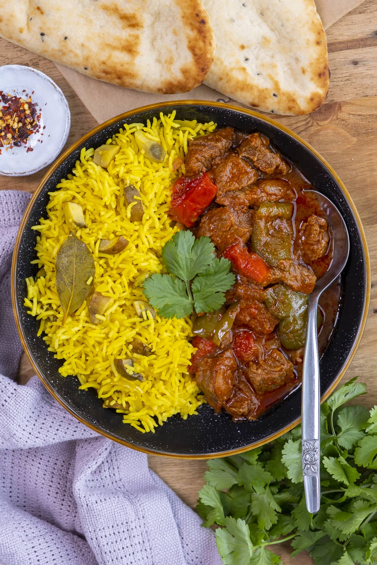 Overhead shot of Easy Peasy Beef Curry on a plate with Mushroom Pilau Rice, surrounded by naans, chilli flakes and coriander