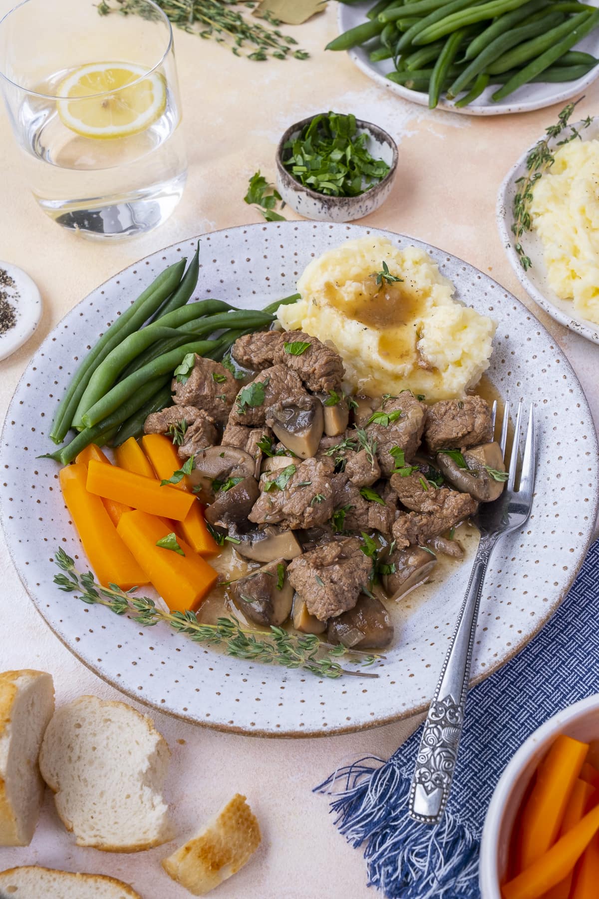 Easy Peasy Beef Stew on a plate with mashed potatoes, carrots and green beans. Surrounded by dishes of veggies and herbs