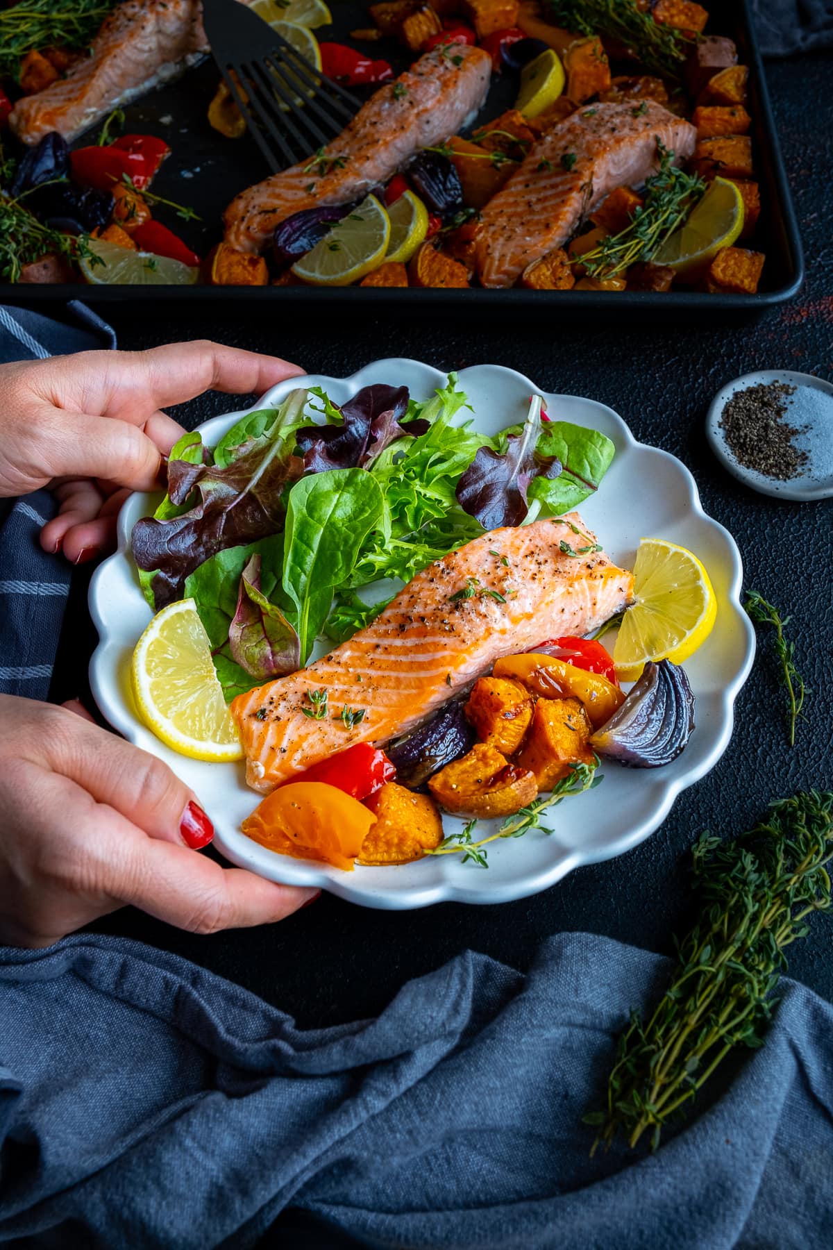 One cooked salmon fillet on a plate with roasted vegetable and salad leaves - a woman's hands are holding the plate