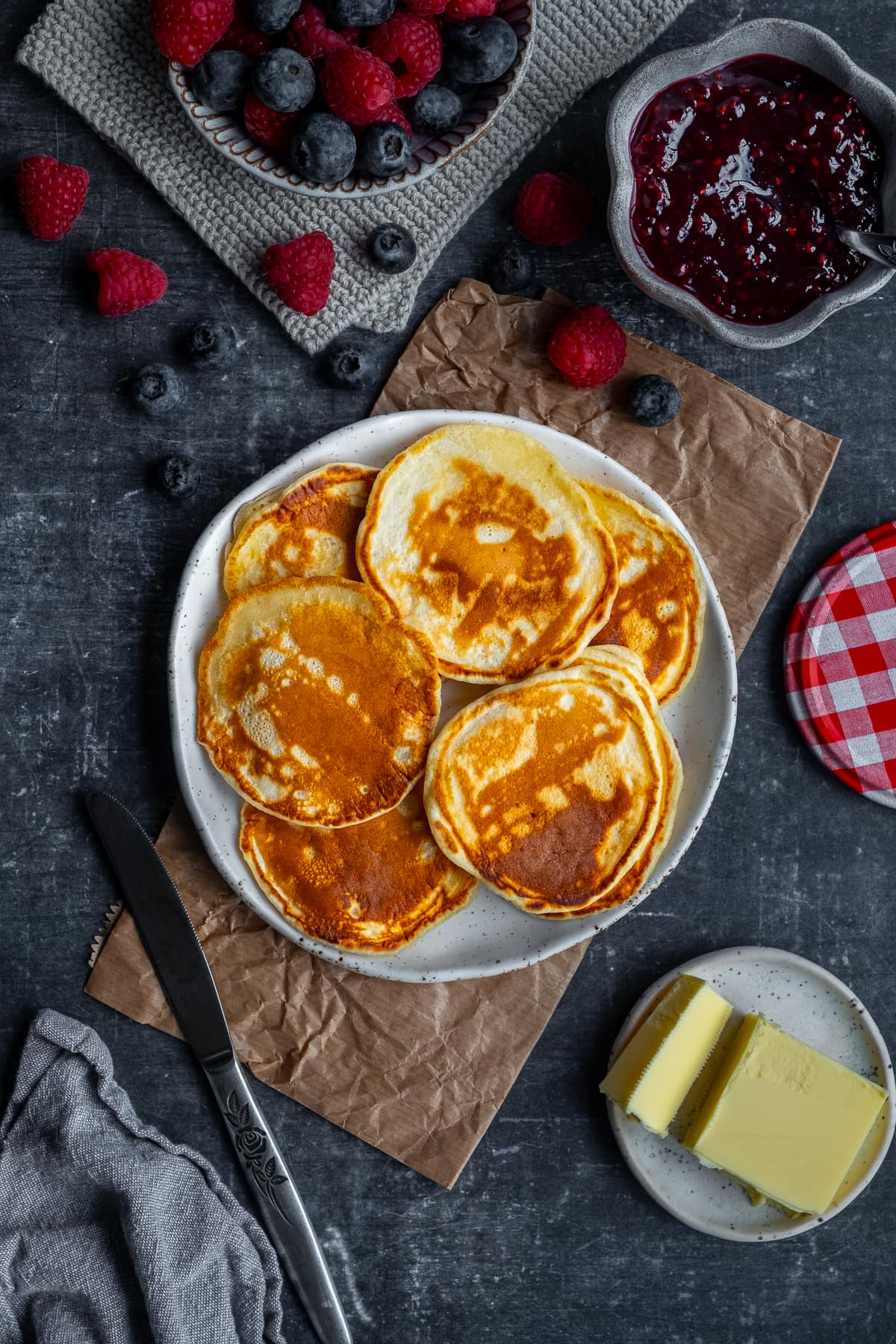 Easy Scotch Pancakes on a plate, surrounded by jam, butter and berries.