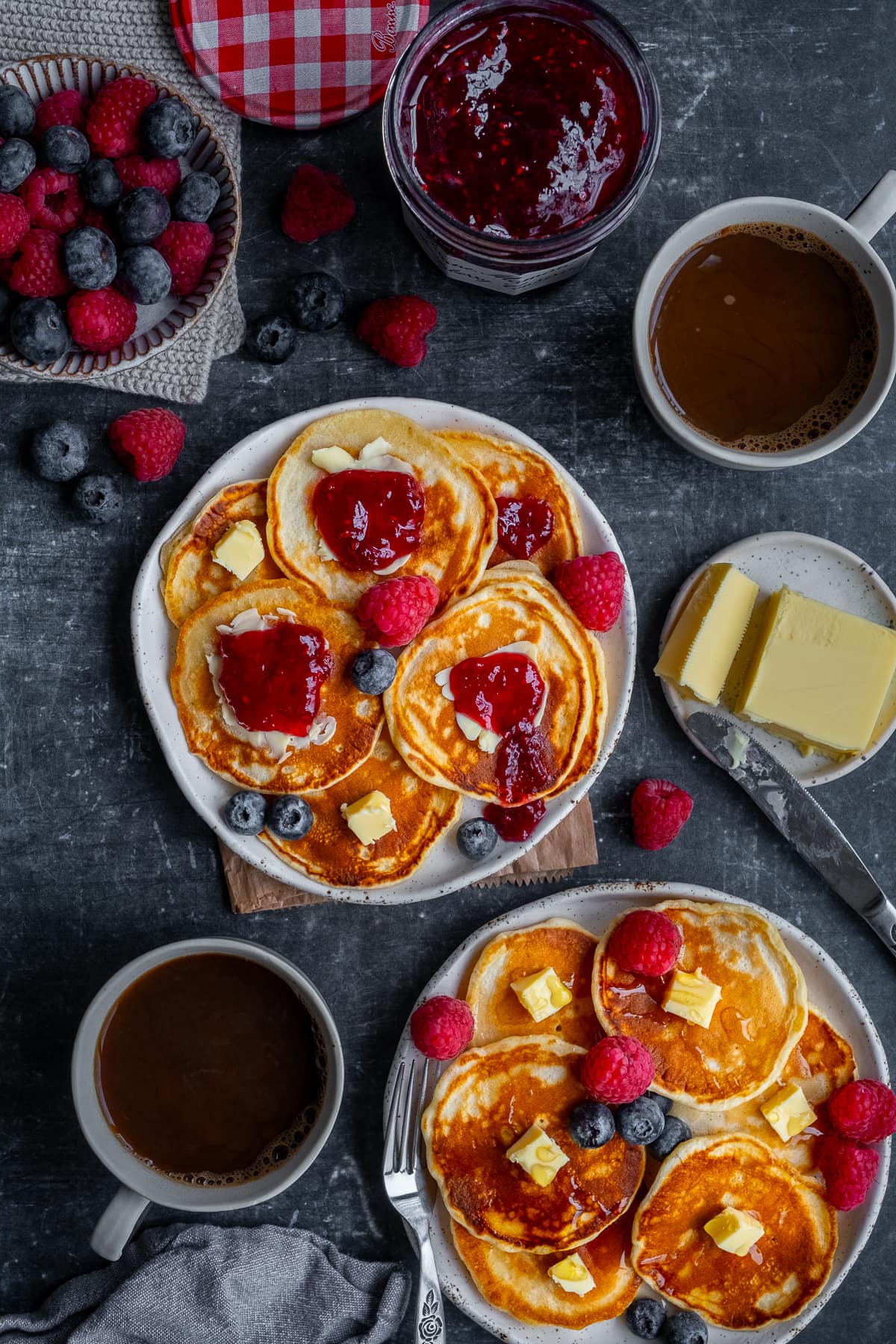 Overhead shot of 2 plates of Scotch Pancakes, served with butter, jam and berries, plus 2 cups of coffee.