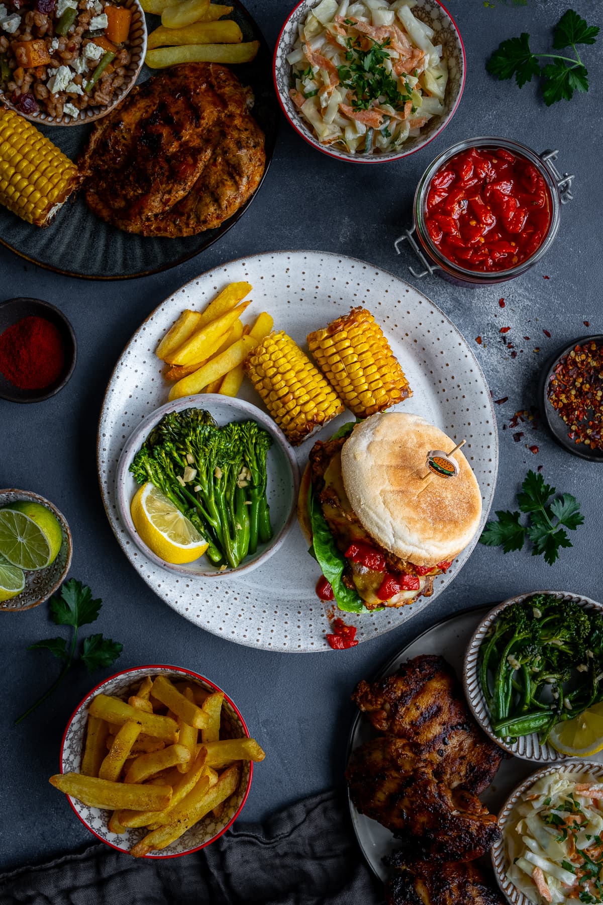 Overhead shot of Homemade Nandos Sunset Burgers surrounded by typical Nandos sides: chips, corn on the cob, Tenderstem Broccoli and Coleslaw