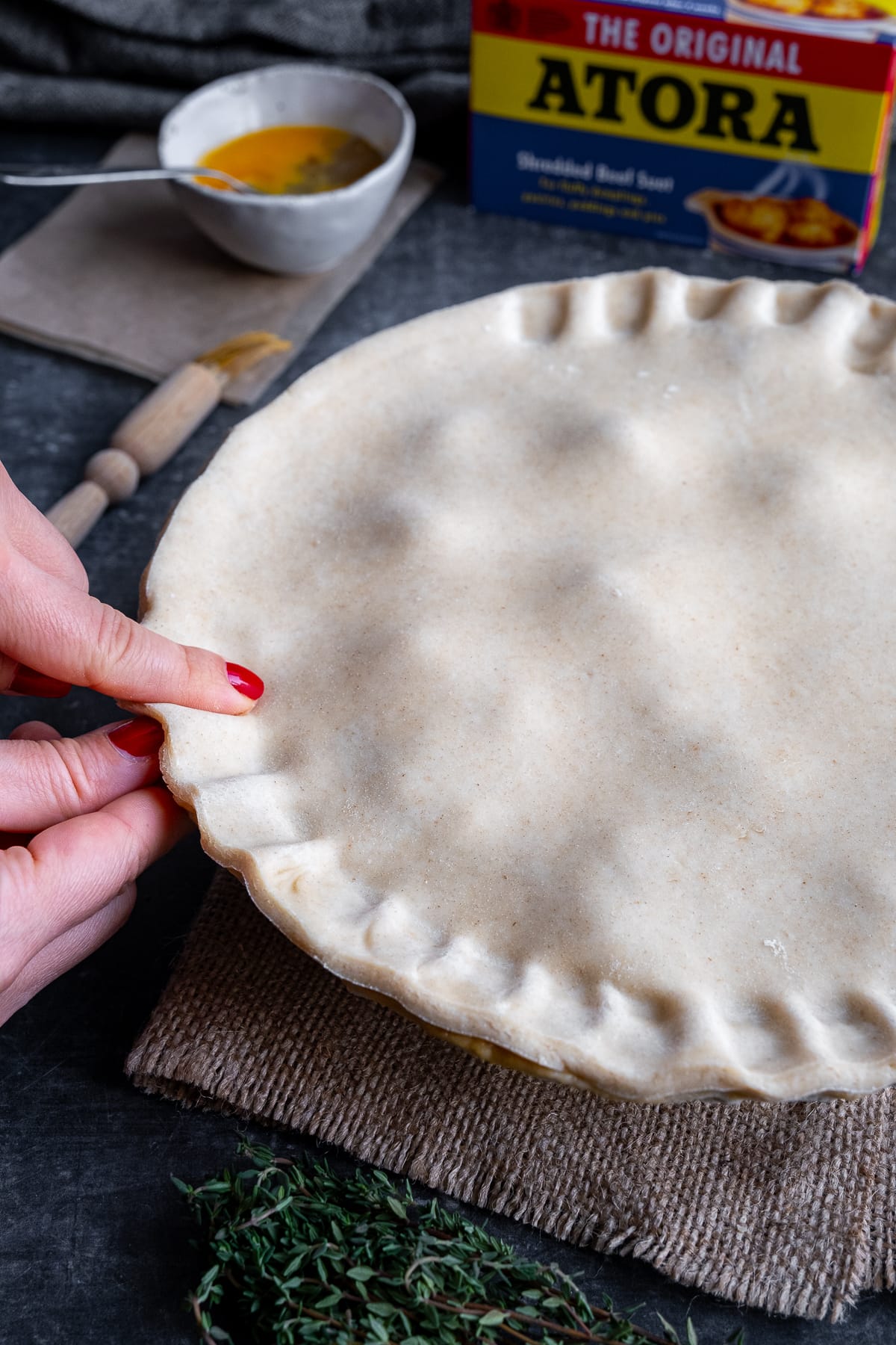 Uncooked suet crust pastry on top of the pie filling. A woman's hands are crimping the edges of the pie.