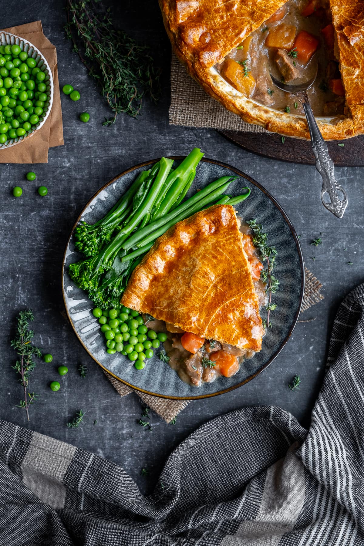 Overhead shot of one portion of Suet Crust Steak Pie on a plate with broccoli and peas.