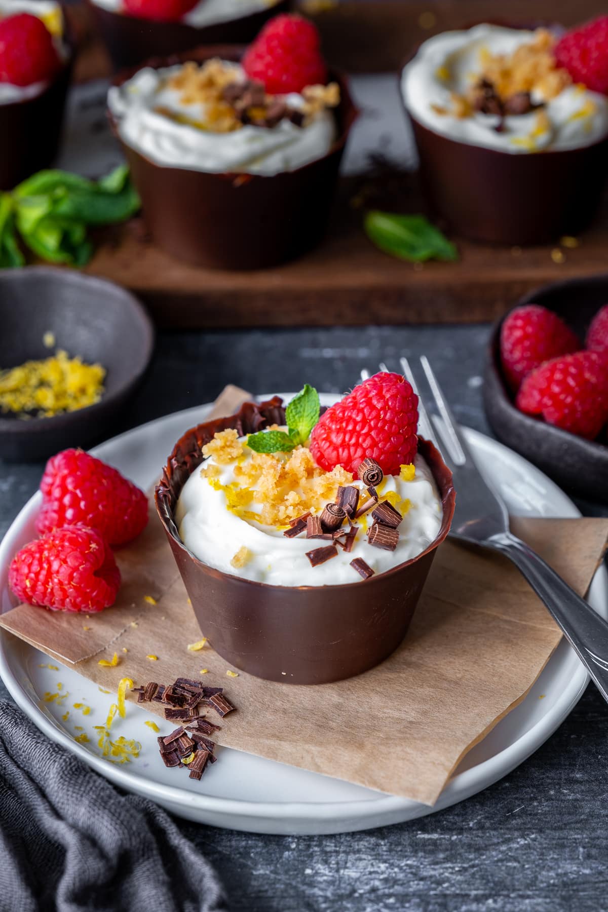 Lemon Cheesecake Chocolate Cups decorated with lemon zest, biscuit crumbs, chocolate sprinkles, fresh raspberries and mint