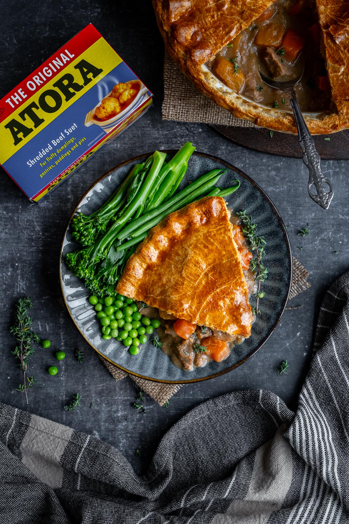 Overhead shot of one portion of Suet Crust Steak Pie on a plate with broccoli and peas.