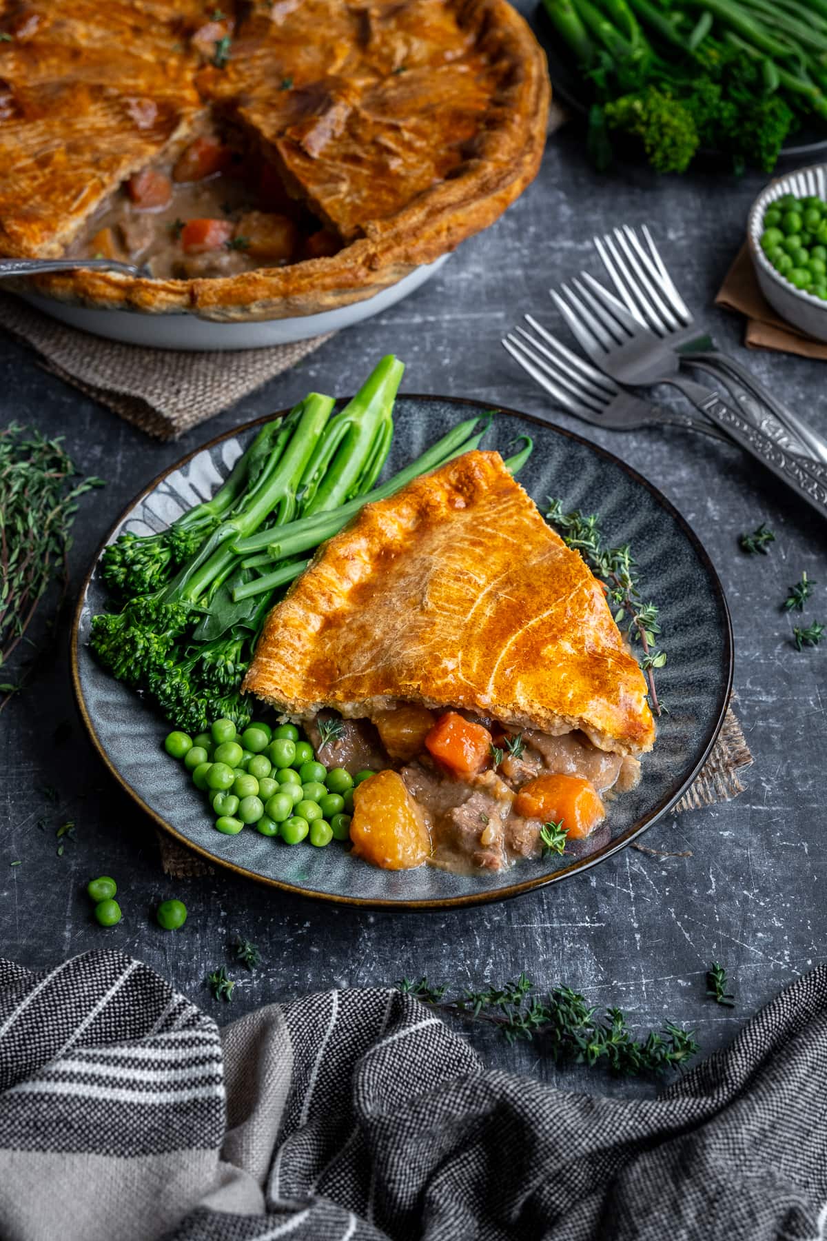 One portion of Suet Crust Steak Pie on a plate with broccoli and peas.